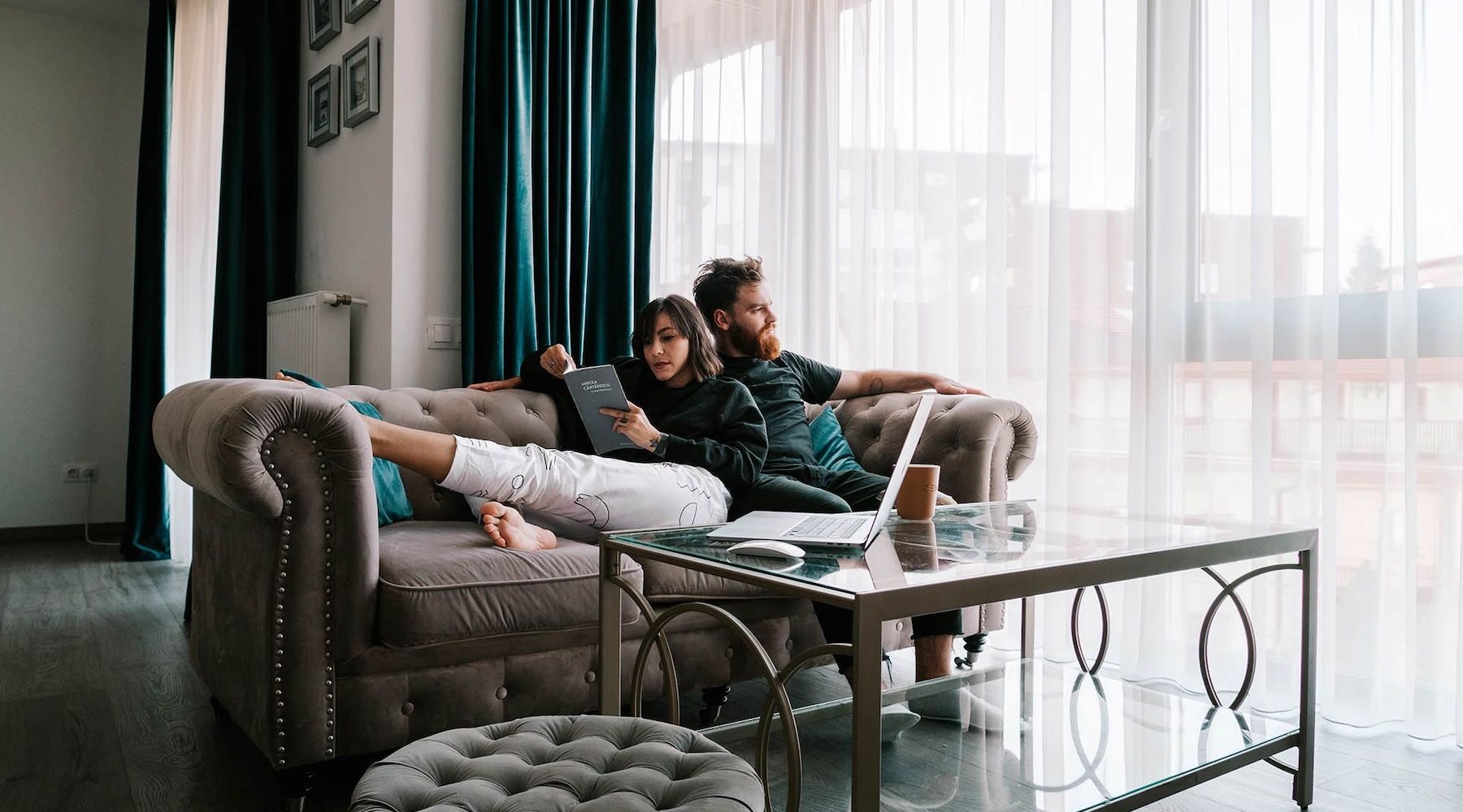 man sits on a couch and looks out the window while his wife lies against him reading a book