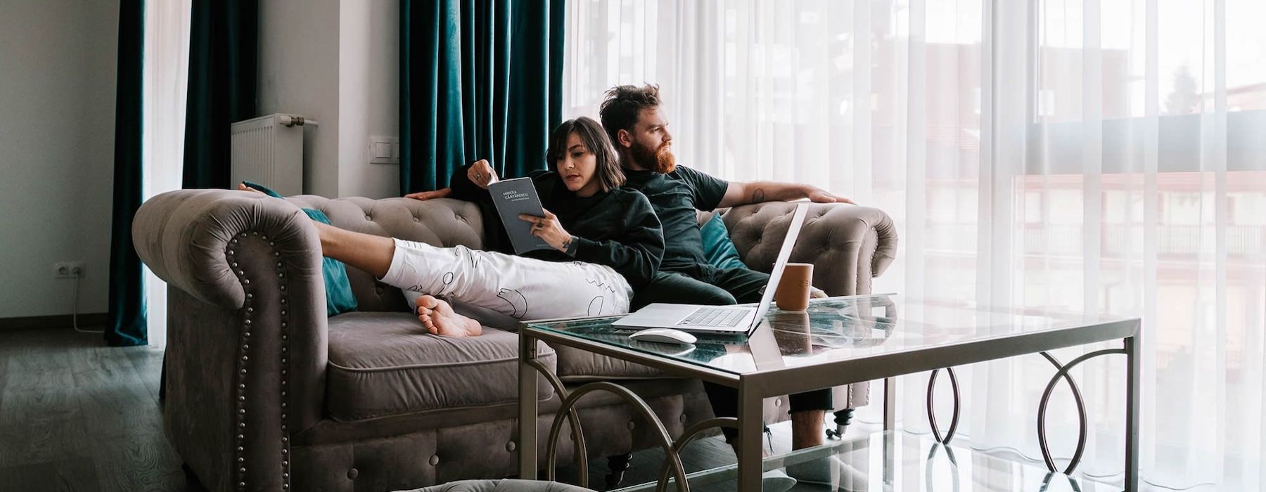 man sits on a couch and looks out the window while his wife lies against him reading a book
